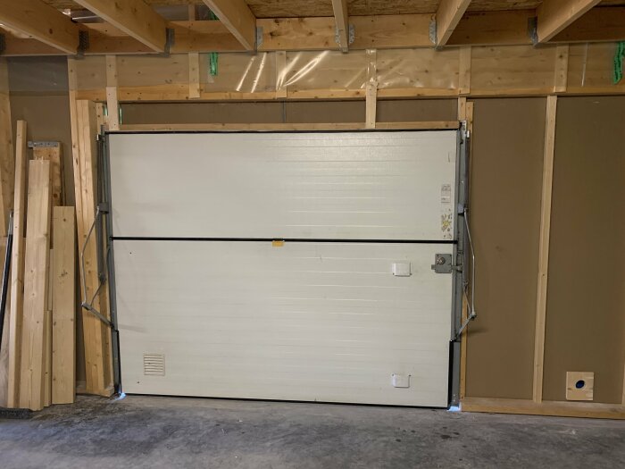 Vit tvådelad garageport monterad i en nybyggd trägaragestomme med oskyddade väggar och takbjälkar.