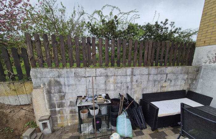 Äldre stödmur av skalblock med synliga skador och uppackade trädgårdsredskap samt möbler framför.