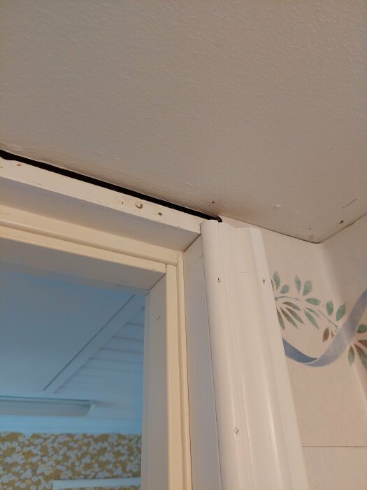 Glipning mellan vitmålad dörrfoder och tak i ett hörn med synliga spikar och tapet.