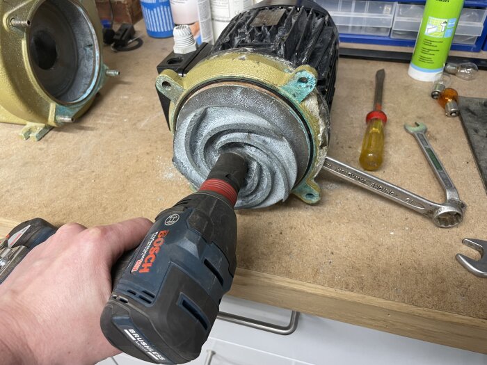 Reparation av elmotor, hand håller borrskruvdragare nära korroderad fläns på bänk med verktyg.