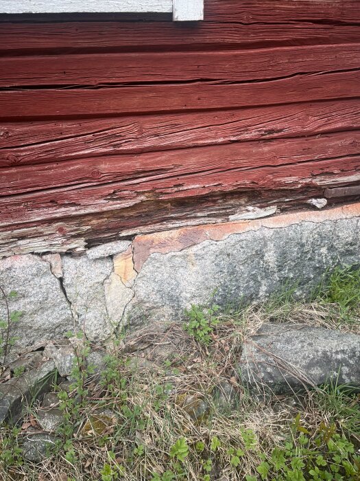 Rödfärgad träpanel på hus med synliga skador och röta nära grunden av sten.