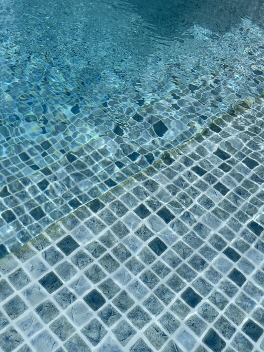 Pool med vattenvågor reflekterar ljus på mosaiktrappor, en gul rand syns på ytan och under vatten.