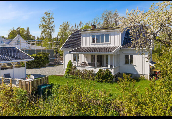Vit villa med veranda och stor betongplatta, belägen på en grönskande höjd under solig himmel.