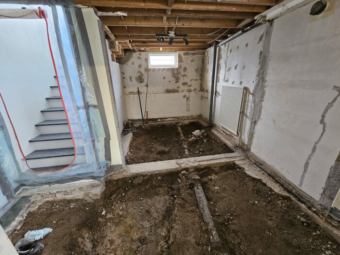 En rå källare under renovering med upptaget golv av cement och synliga avloppsrör, nära en trappa och fönster.