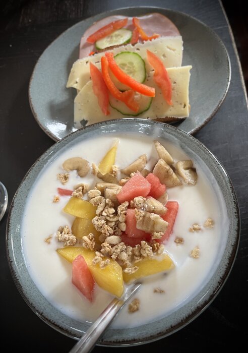 Frukostmål med yoghurt, färsk frukt och müsli samt en smörgås med ost, skinka, gurka och paprika.