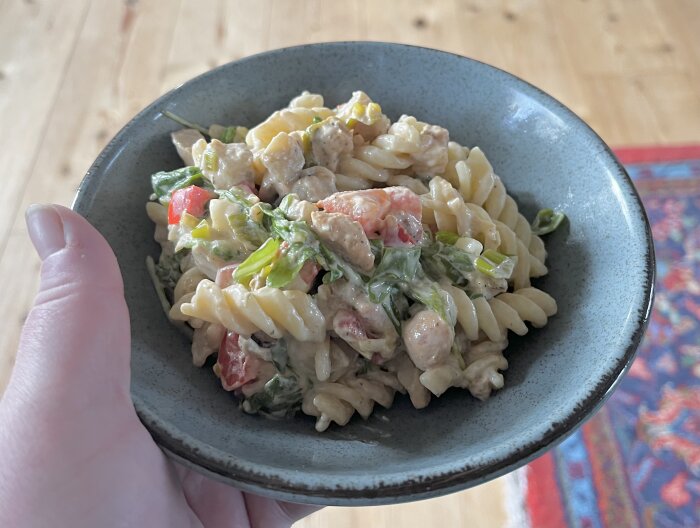 Hand håller en skål med pastasås, innehållande kyckling, salladslök, tomat, paprika, aubergine och sallad.