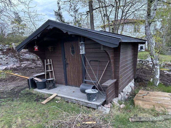 Brun trästuga med svart tak, dörr och några verktyg framför, storlek 4,20 x 4,40 meter.