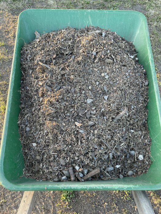 Översta lagret av en kompost med småsten, bark och torra växtdelar i en grön skottkärra, indikerar problem med jordens kvalitet.