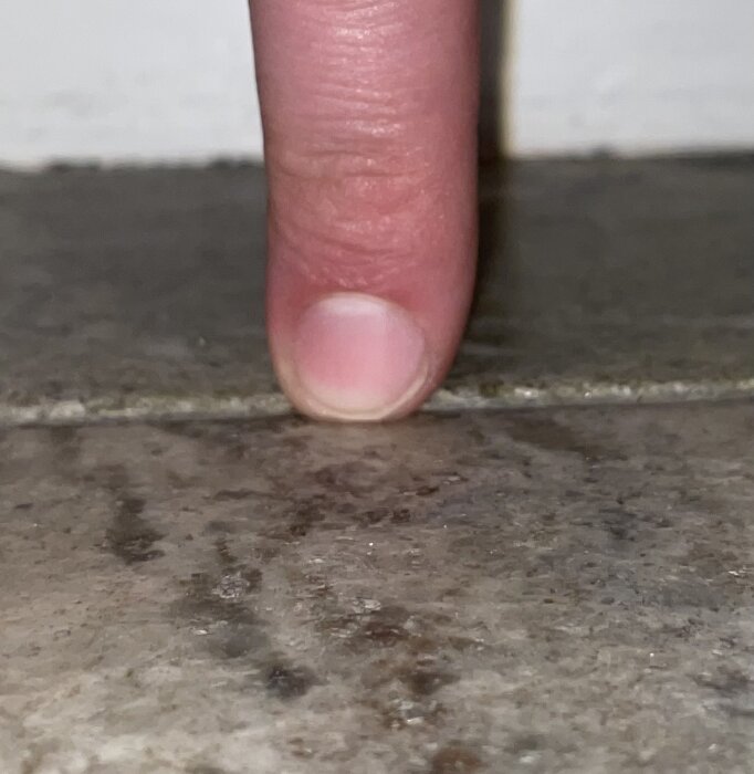 Närbild av ett finger som pekar på en ojämn skarv mellan stenplattor på ett stengolv.