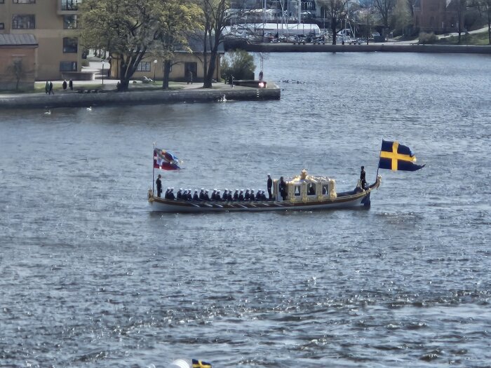 Traditionell svensk roddbåt med besättning och flagga på vattnet.