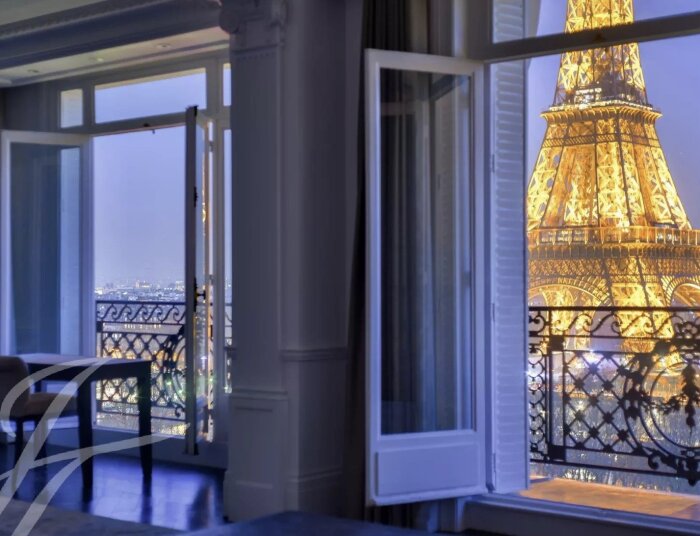 Utsikt från ett fönster med öppen balkongdörr visar Eiffeltornet upplyst om natten.