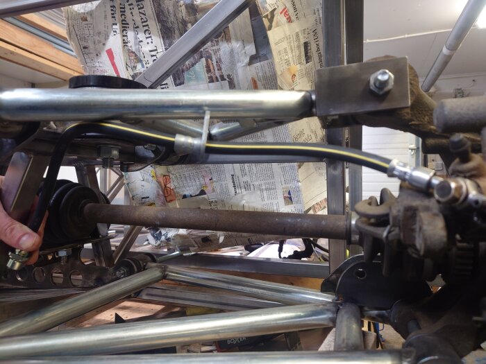 Del av bilbygge med en hand som justerar bromsslang nära hjulupphängningen, metallrör och bakgrund av tidningar.
