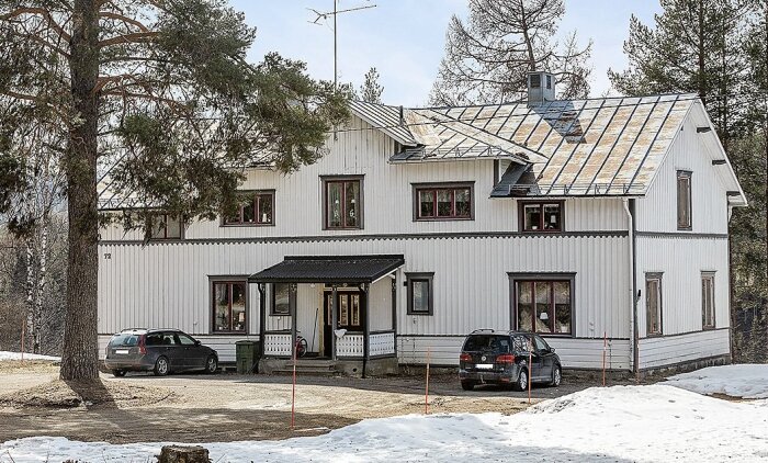 Vitt hyreshus med solpaneler på östlig taknock, röda fönsterluckor, omgivet av höga träd, med två bilar parkerade framför.