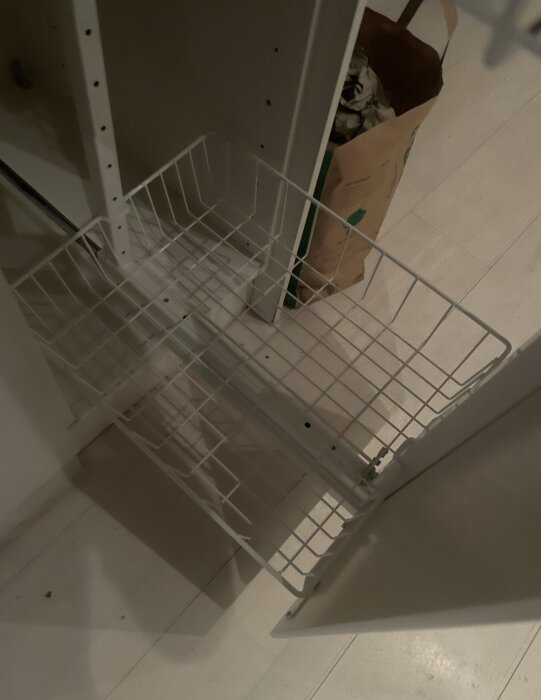 Vit trådkorg i öppet köksskåp med en papperskasse i bakgrunden.