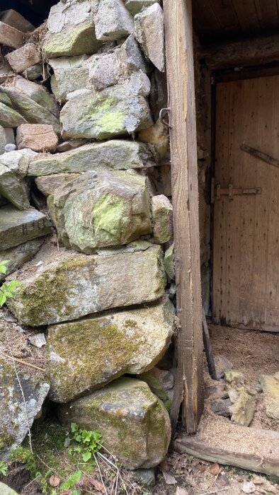 Stenmur vid jordkällare med lossnade stenar och en sprucken träkarm runt en stängd dörr.