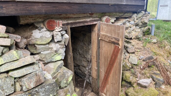 Skadad vägg vid jordkällardörr med spruckna stenar och dörrkarm under en stuga.