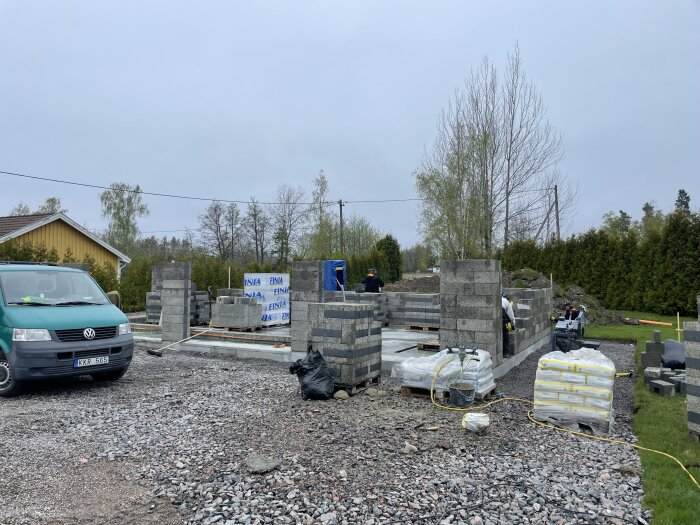 Byggplats med nyuppsatta gråa betongblock till en grund, byggmaterial i förgrunden och arbetare i bakgrunden.