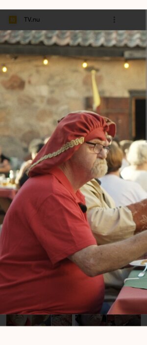 Man i röd skjorta och röd hatt med guldkant sitter vid ett bord utomhus.