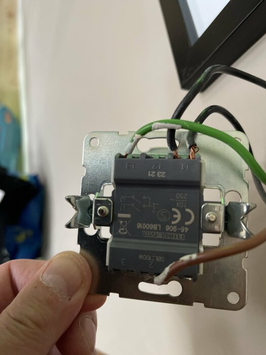 Hand håller en elektrisk väggströmbrytare med exponerad kabelanslutning under installation.