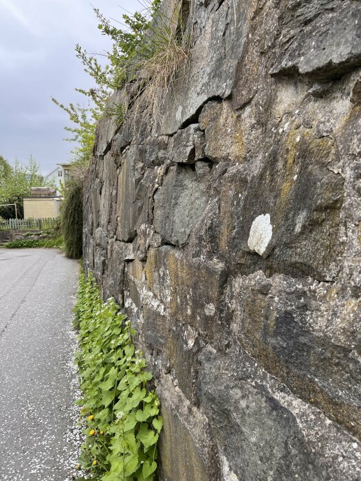 Stenmur med vittrat bruk och lösa stenar längs en bilväg med grönska.