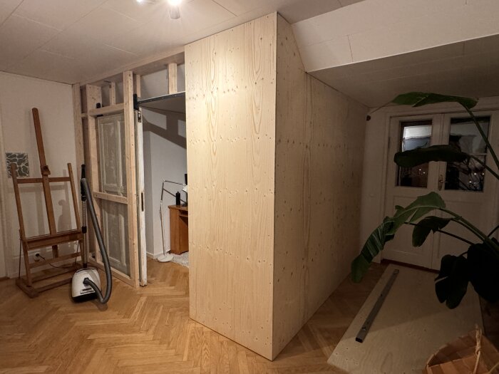 Nybyggt träregelverk beklätt med plywood i ett gammalt rum med växter och målarutrustning.