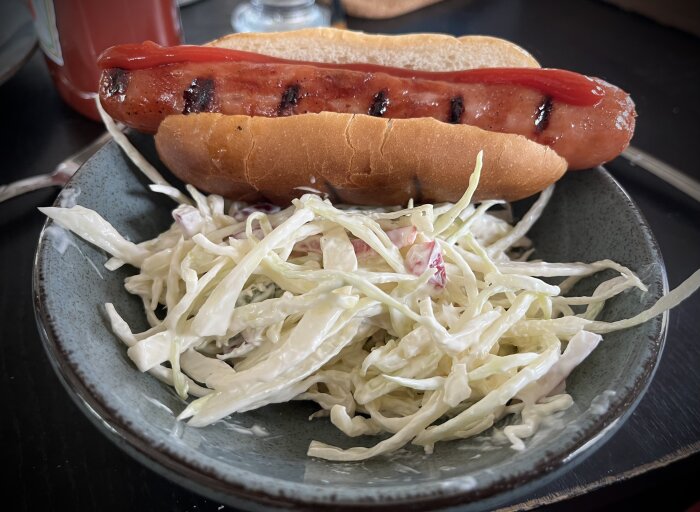 Grillad ost- och baconkorv med ketchup i bröd och en skål coleslaw.