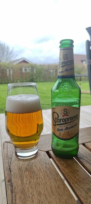 Ett glas med ljusgul öl och en grön Staropramen alkoholfri ölflaska på ett träbord utomhus.