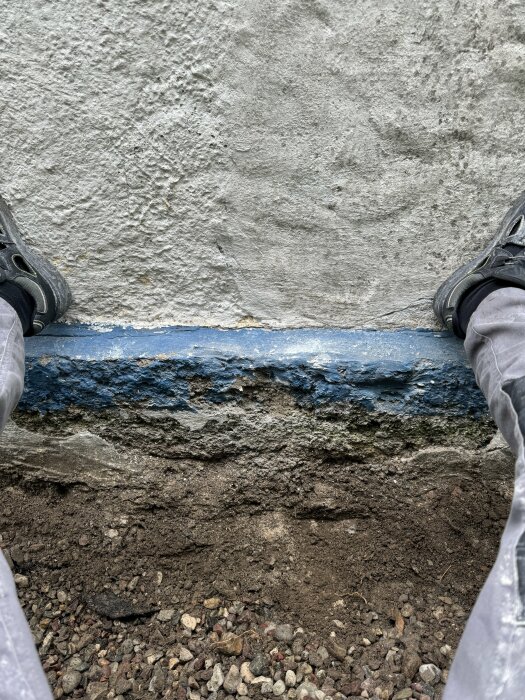 Vy neråt mot en vittrande grund med synlig blå väv och jord, skor syns i bildens nedre kant.