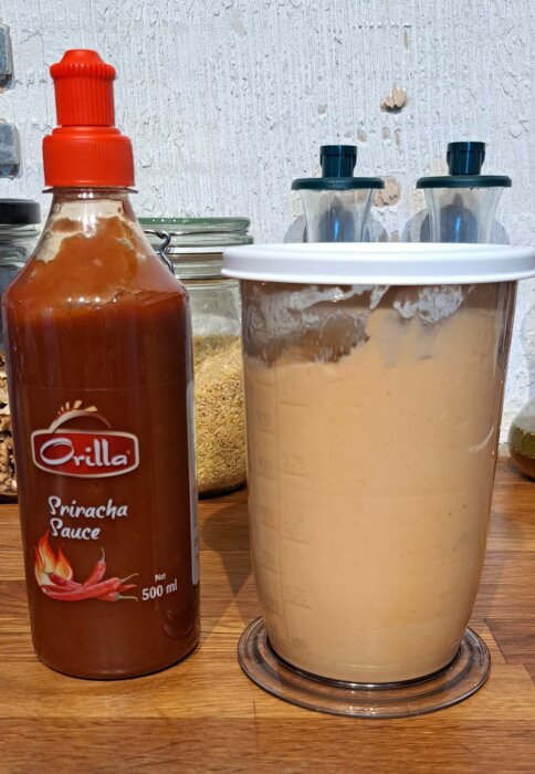 Flaska med sriracha sås bredvid behållare med hemgjord sriracha majonäs på köksbänk.
