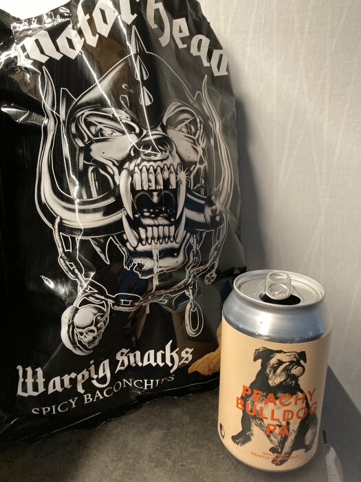 En öppnad burk av Peachy Bulldog PA öl bredvid en påse Warpig Snacks Spicy Baconchips med Motorhead-tema.