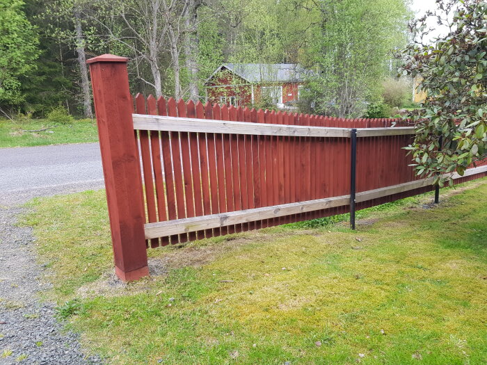 Nymålat rödbrunt staket vid en tomt med gräs och träd i bakgrunden.