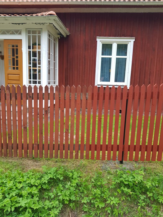 Delvis nymålat rött staket framför en röd stuga med vit dörr och fönster.
