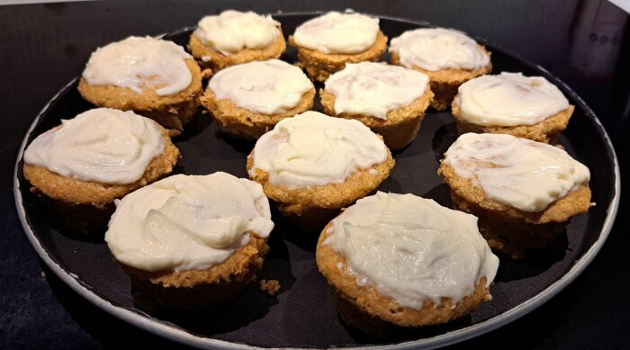 Nybakade morotsmuffins med frostingtäcke på en mörk bakplåt.