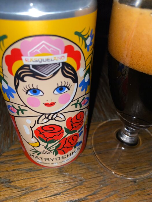 Färgglad burk av Spansk Imperial Stout med Matryoshka-dockdesign, delvis fylld ölglas med mörkt Stout syns till höger.