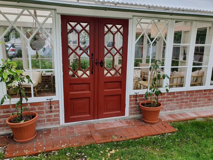 Röda pardörrar till ett växthus med vita fönster, tegelstenar och krukväxter framför.