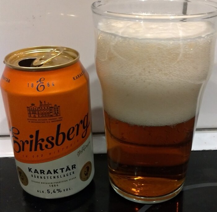 Öppnad ölburk av märket Griksberg och ett glas fyllt med lageröl, stående på en bordsskiva.