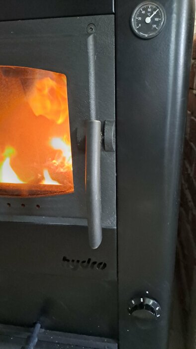 Närbild av en tänd köksspis med eldstad och synliga lågor bakom glaset, handtag och temperaturmätare.