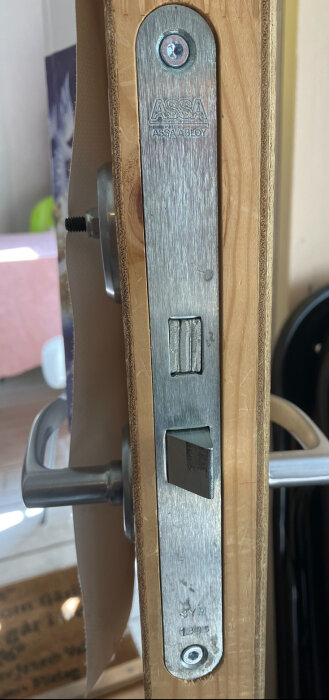Ett dörrhandtag med en synlig låsmekanism märkt "ASSA ABLOY" monterat på en ofullständigt stängd dörr.
