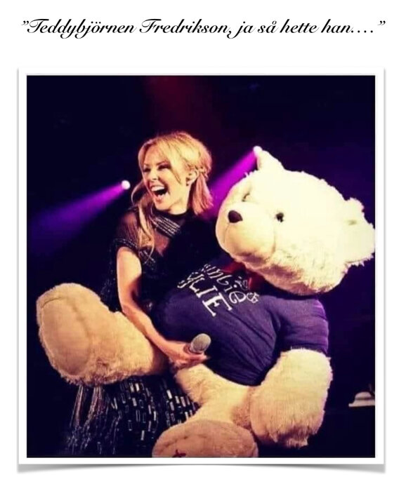 Sångerska skrattande med mikrofon bredvid en stor teddybjörn på scen.