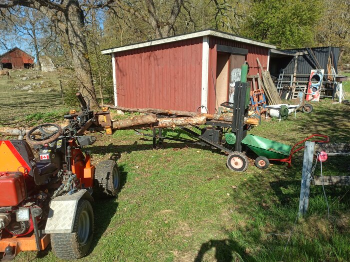 Traktor med vedklippare lastad med granar vid röd vedbod på en gård.