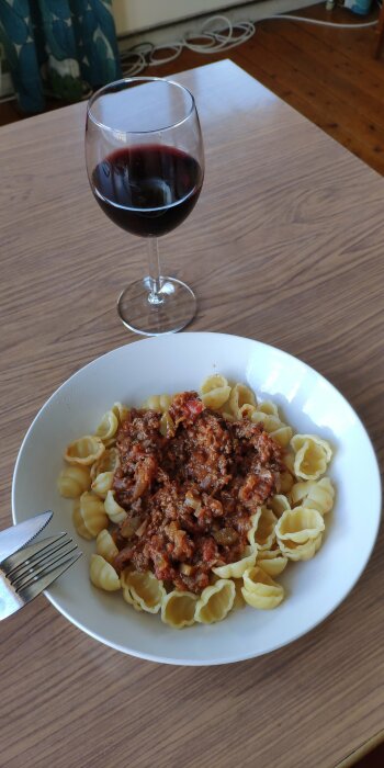 Tallrik med bolognese på pastaskal och ett glas rött vin, uppdukat på ett träbord.