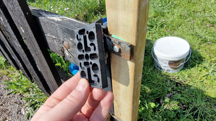 Hand håller shims bredvid en trästolpe i början av en staketbyggnation med gammalt staket och hink med träolja i bakgrunden.