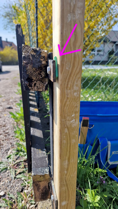 En tryckimpregnerad stolpe fästad mot ett järnstaket med pallning och skruv, markerat med en rosa pil.