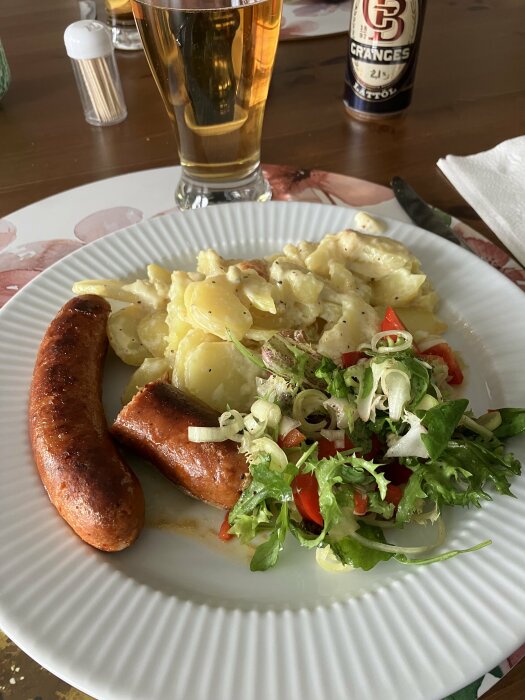 Chorizo med potatisgratäng och sallad på vit tallrik, med öl i glas och kryddburk i bakgrunden.