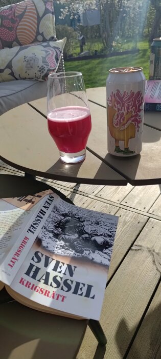Rosa öl i glas bredvid burk med Berry Lemonade-design på soligt utebord med bok.