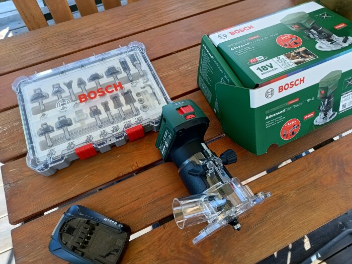 Bosch 18V handöverfräs med batteri, låda med frässtål och produktkartong på ett träbord.