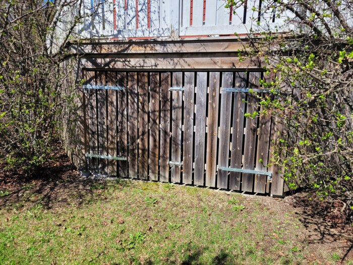 Träskjul med två inbyggda dörrar, visuellt kamouflerade som del av den omgivande träpanelen, mot en bakgrund av grönskande buskar.