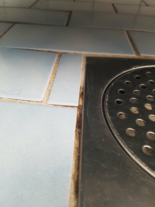 Närbild på badrumsgolv med blåa klinkers och svarta kakelfogar intill en duschavloppssil.