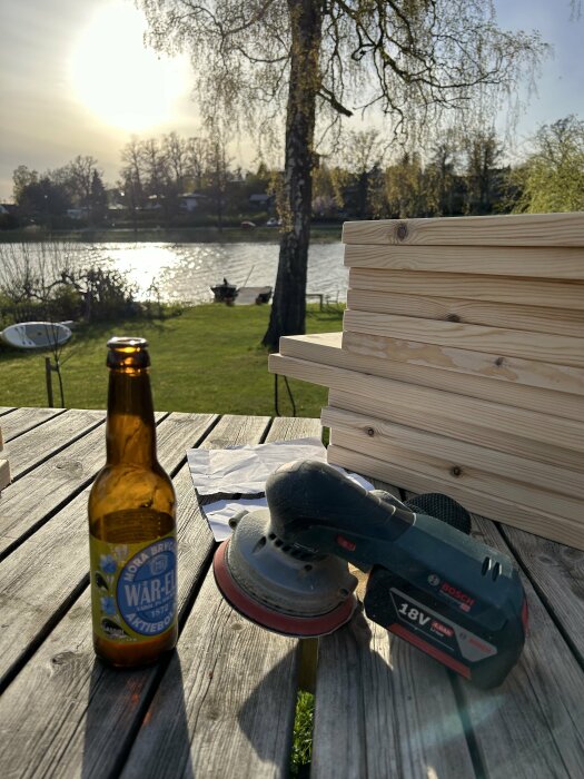 Batteridriven slipmaskin och en ölflaska på ett träbord med staplade träplankor i bakgrunden, utomhus vid sjö.