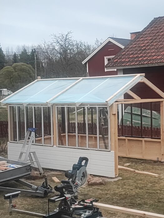 Uppbyggnad av ett växthus med genomskinlig plasttak och vita väggar vid ett hus, verktyg syns framför.